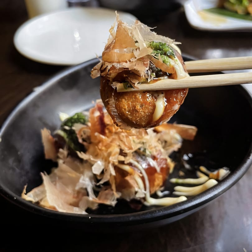 Chopsticks holding a takoyaki ball topped with bonito flakes, sauce, and green onions over a black bowl.
