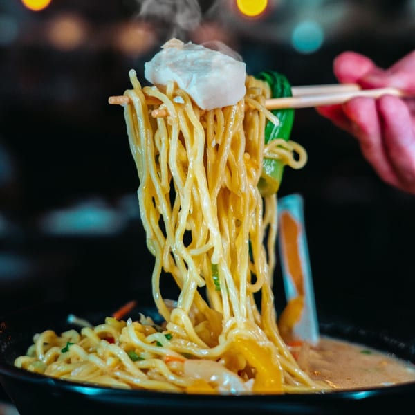 Steaming ramen noodles with pork belly and greens being lifted by chopsticks from a black bowl.