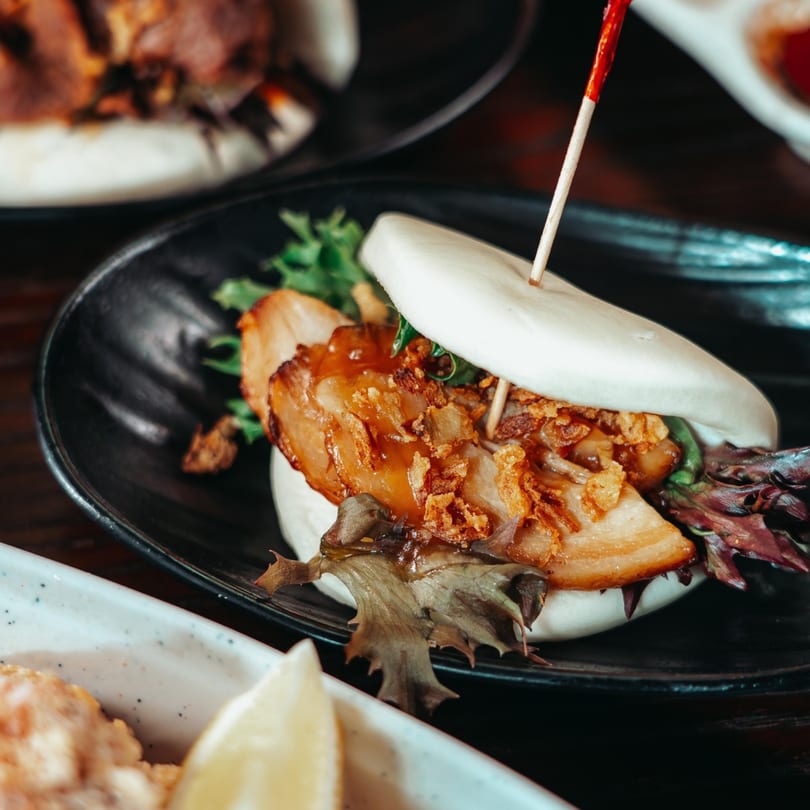 A close-up of a bao bun filled with pork belly, lettuce, and crispy fried onions.