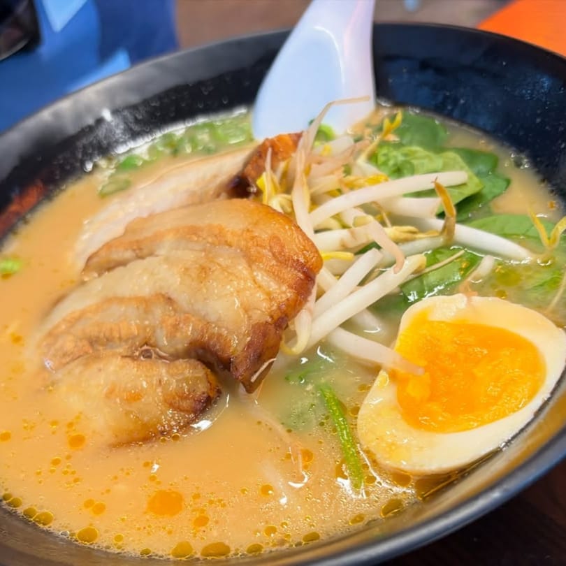Close-up of ramen with sliced pork, bean sprouts, spinach, and a soft-boiled egg in a black bowl.